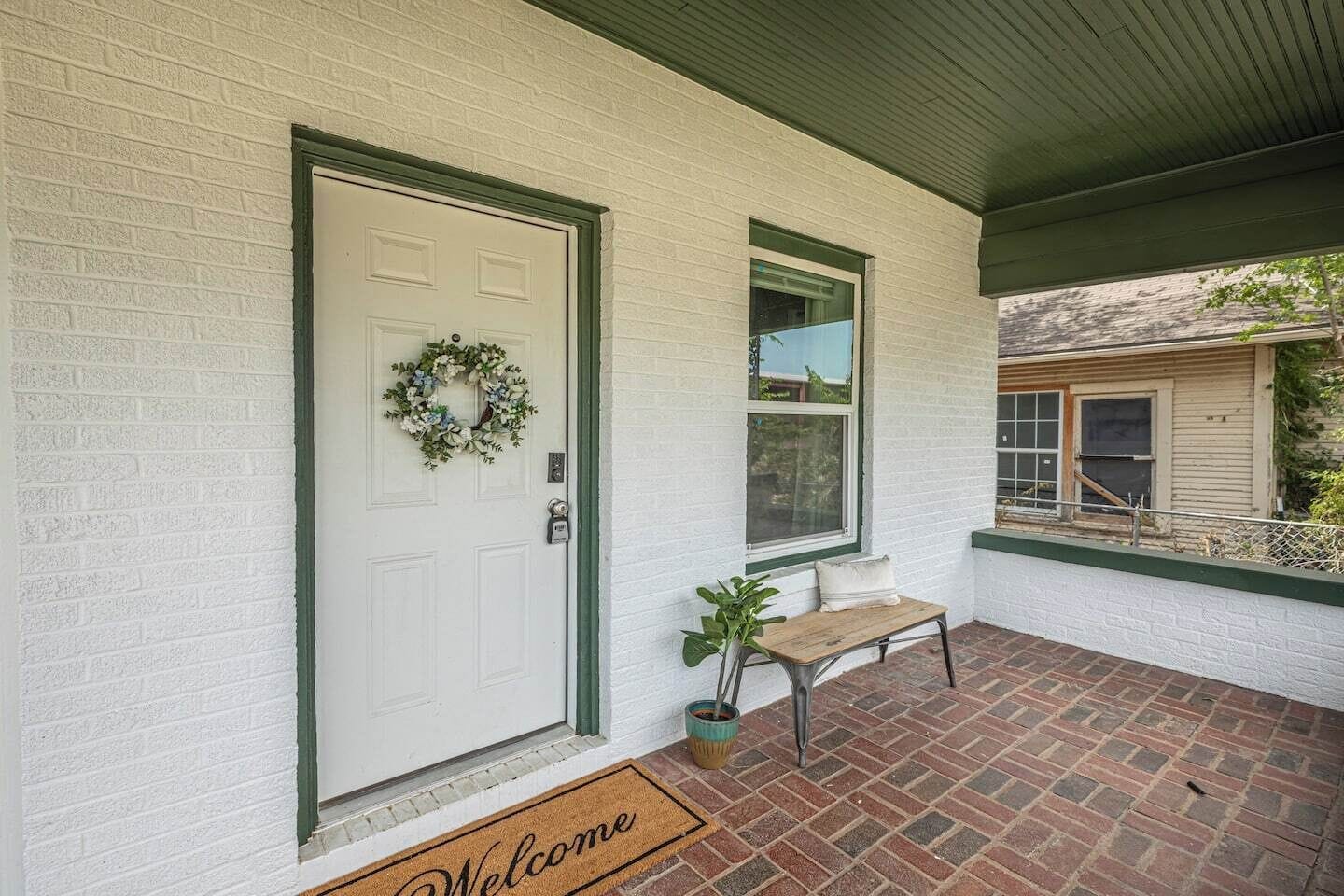 A white door with a wreath, a bench, and a welcome mat outside a building with a brick floor and a green roof.