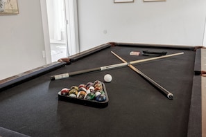 Une table de billard avec des billes et des queues, une fenêtre avec des rideaux et un tableau encadré au mur.