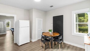 A modern kitchen with a white refrigerator, a wooden dining table with four chairs, and a window with a view of greenery. - Modern 2BR Home with Pool Table & Yard (Abilene)