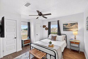 Une chambre à coucher comprenant un lit, des tables de chevet, un ventilateur de plafond, une fenêtre avec des rideaux et une vue sur l’extérieur.