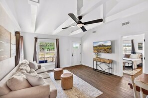 Un salon moderne avec un canapé, des poufs, une table basse, un ventilateur de plafond et un tableau accroché au mur.