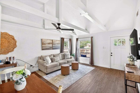 Un salon spacieux avec un plafond haut, un ventilateur de plafond, une grande fenêtre avec des rideaux, un canapé, une table basse et un téléviseur posé sur un meuble.