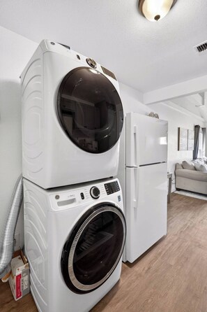 A compact laundry room with a stacked washer and dryer, a refrigerator, and a wooden floor. - Peaceful 2BR Home with Modern Comforts (Abilene)