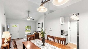 Uma sala de jantar com mesa de madeira, cadeiras, um vaso com plantas, ventilador de teto e luminárias pendentes.