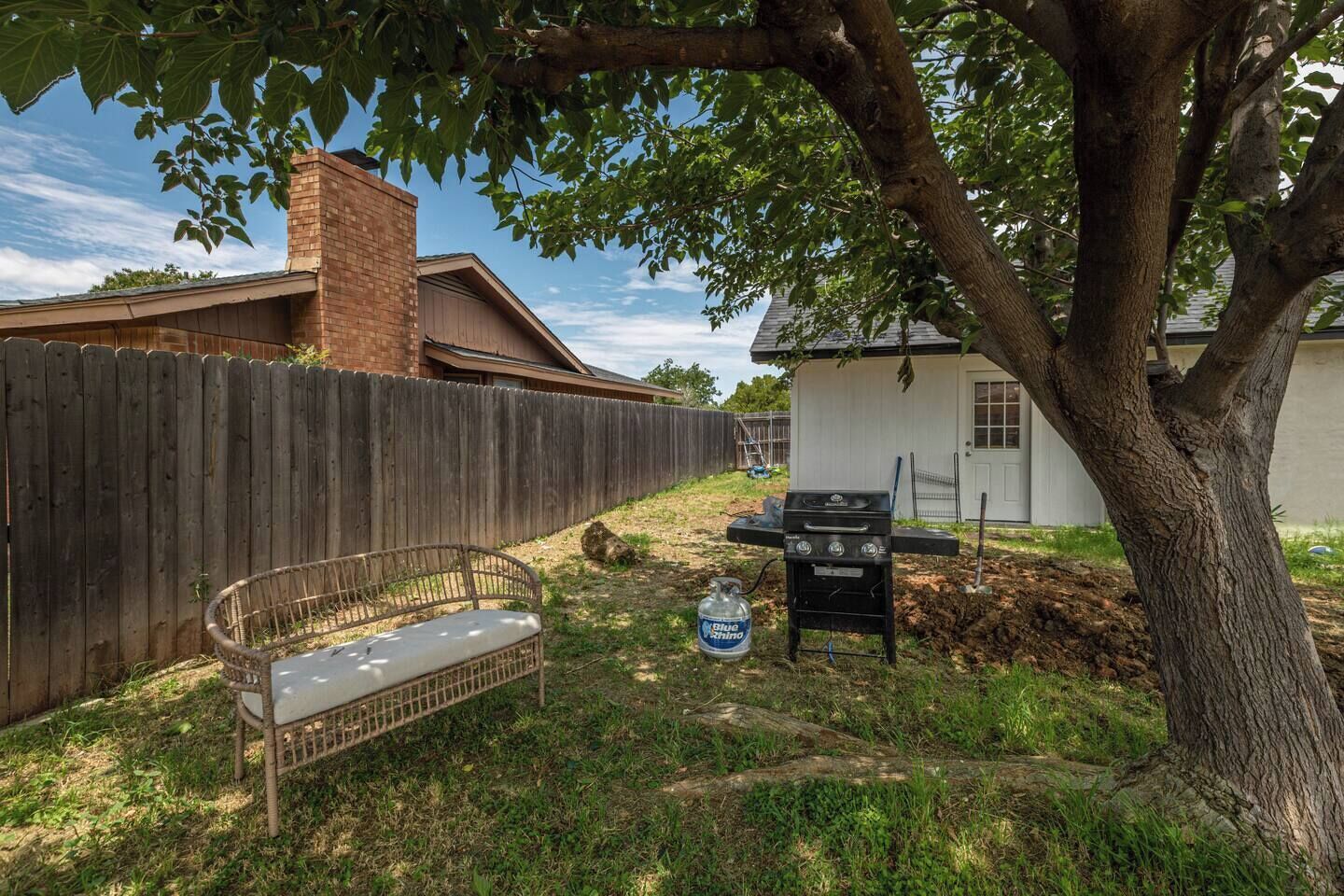 Ein Garten mit Holzzaun, Grill, Korbsessel und Baum.