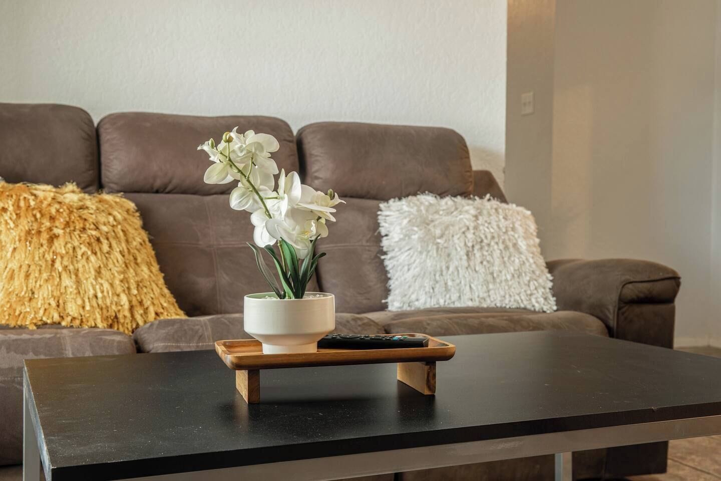 Un salon avec un canapé marron, un coussin blanc et un coussin jaune moelleux, un petit vase blanc contenant des fleurs blanches, et une télécommande posée sur un plateau en bois.
