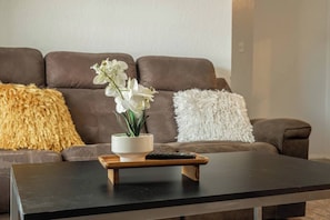 Un salon avec un canapé marron, un coussin blanc et un coussin jaune moelleux, un petit vase blanc contenant des fleurs blanches, et une télécommande posée sur un plateau en bois.