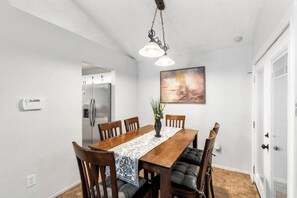 Un coin repas comprenant une table en bois, des chaises, une suspension, un tableau et un réfrigérateur.