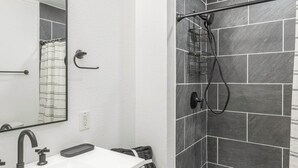 Casa de banho com toucador branco, espelho, chuveiro com porta de vidro e tapete de banho preto.