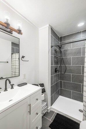 Ein Badezimmer mit weißem Waschtisch, einem Spiegel, einer Dusche mit Glastür und einer schwarzen Fußmatte.
