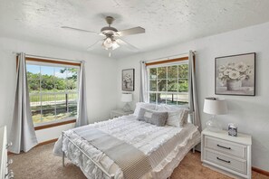 Une chambre à coucher comprenant un lit, deux tables de chevet, un ventilateur de plafond et deux fenêtres avec des rideaux.