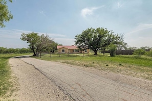 A gravel driveway leading to a house with a red roof, surrounded by trees and a fenced yard. - Country Living: 3BR Hot Tub Escape: Close to all! (Abilene)