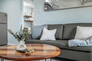 Een woonkamer met een grijze bank, een ronde houten salontafel, een witte vaas met groen en een afstandsbediening.