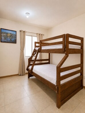 Een stapelbed met houten frame en wit beddengoed in een kamer met een betegelde vloer en een raam met gordijnen.