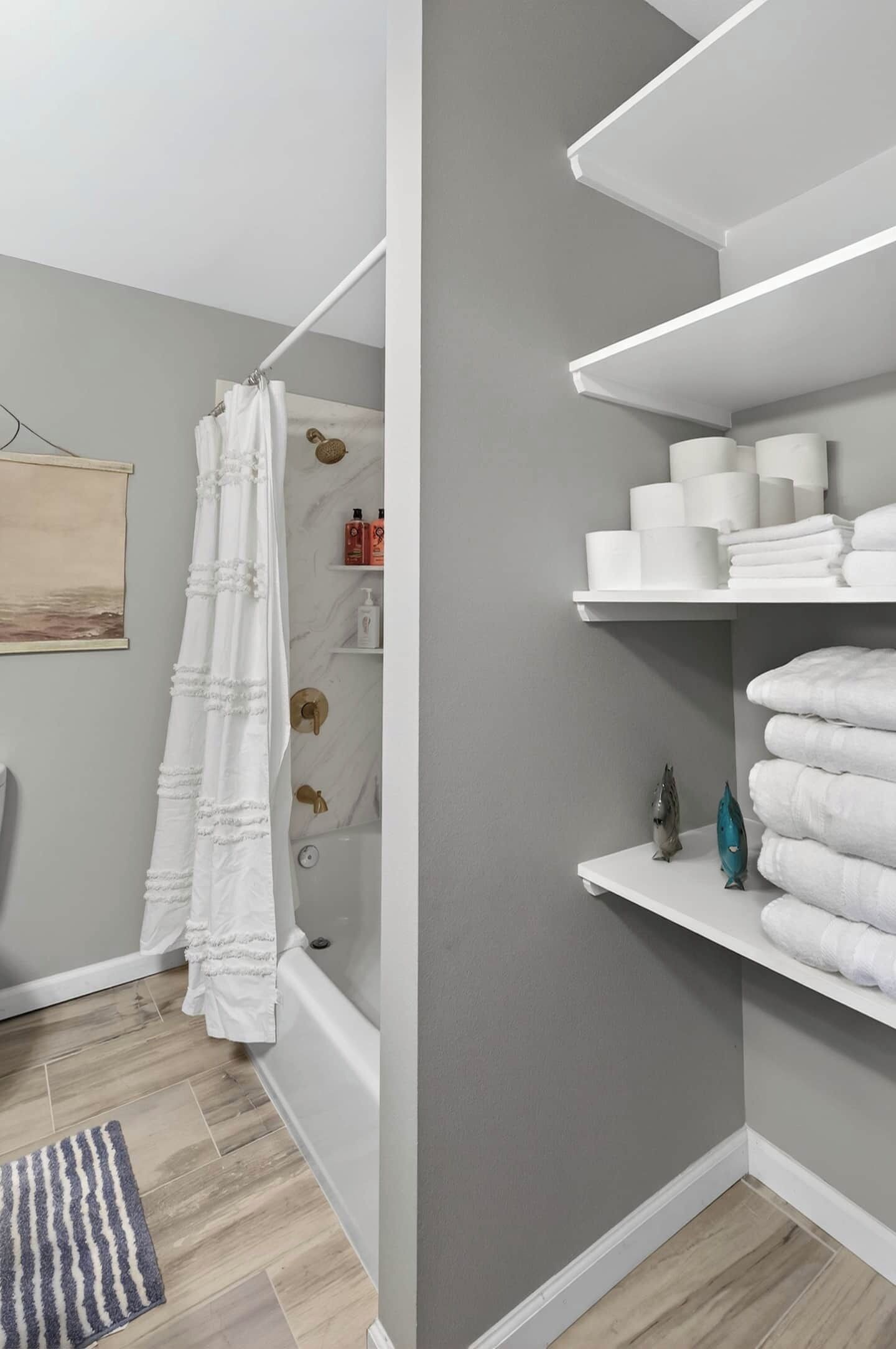 A bathroom with a shower curtain, a bathtub, a towel rack, and shelves with toiletries.