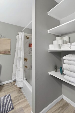 Un baño con cortina de ducha, bañera, toallero y estantes con artículos de aseo.
