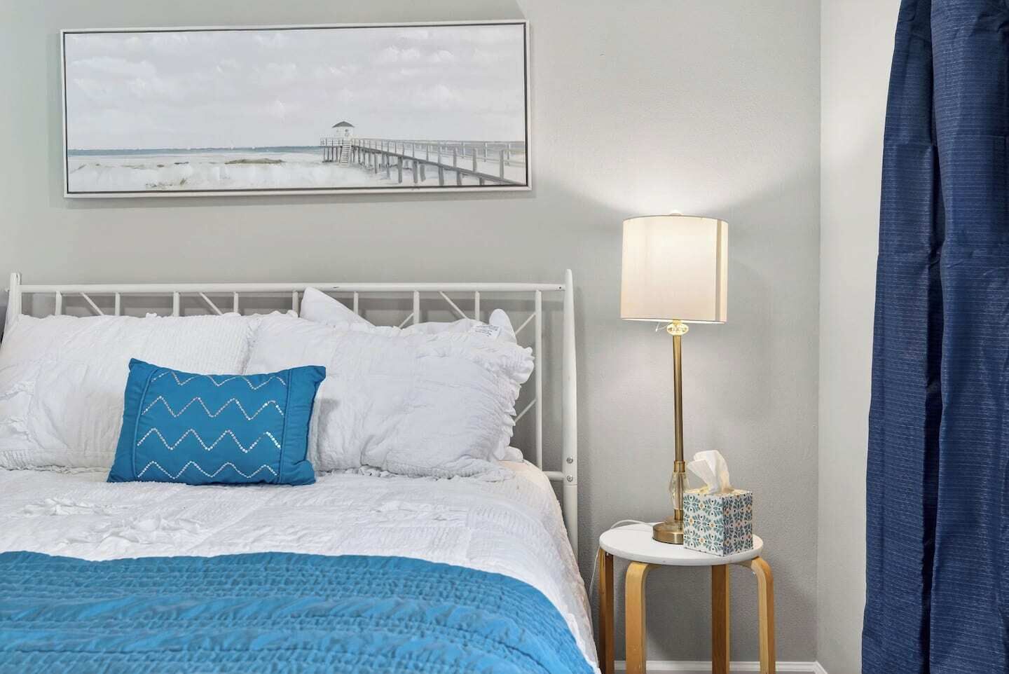 A bedroom with a bed, a nightstand with a lamp, a picture on the wall, and a window with curtains.