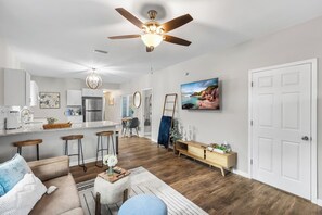A modern kitchen with a central island, a dining area with a wooden table, a living room with a sofa and a coffee table, and a hallway with a painting and a door.