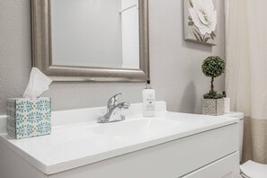 Une salle de bain avec un lavabo blanc, un miroir, un distributeur de savon liquide, une boîte de mouchoirs et une petite plante en pot.