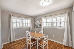 Un coin repas avec une table en bois et des chaises blanches, deux fenêtres avec des rideaux blancs et un luminaire au plafond.