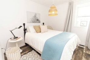 A bedroom with a bed, a bedside lamp, a wall-mounted picture, a window with curtains, and a hanging light fixture. - Best Location in Wellington, Prince Edward County (Prince Edward)