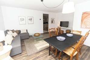A modern living room with a grey sofa, a black dining table, and wooden chairs. There is a television on a stand, a painting on the wall, and a small table with a bowl. - Best Location in Wellington, Prince Edward County (Prince Edward)