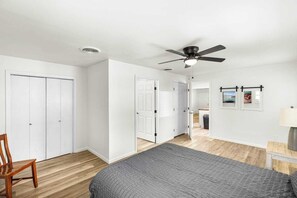 A bedroom with a bed, a ceiling fan, a wooden chair, and a hallway leading to a bathroom. - Modern 3BR Home with Custom Wood Counters (Abilene)