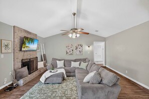 Ein Wohnzimmer mit einem grauen Ecksofa, einem Kamin, einem Deckenventilator und einem an der Ziegelwand befestigten Fernseher.