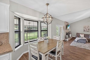 A dining area with a wooden table and chairs, large windows, a chandelier, and a living room with a sofa and television. - Cozy & Modern 3BR Home with Huge Backyard (Abilene)