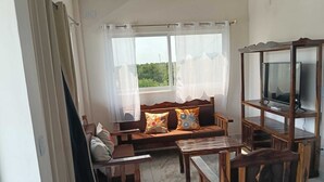 Ein Zimmer mit einer Couch, einem Holz-Fernsehschrank und einem Fenster mit durchsichtigen Vorhängen.