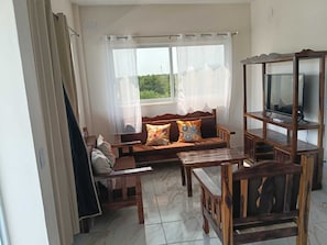 Ein Zimmer mit einer Couch, einem Holz-Fernsehschrank und einem Fenster mit durchsichtigen Vorhängen.