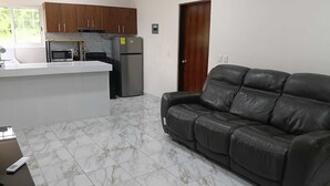 A living room with a leather sofa, a kitchen area with a refrigerator and stove, and a wooden door.