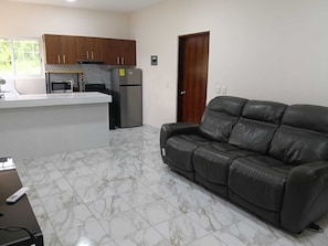 A living room with a leather sofa, a kitchen area with a refrigerator and stove, and a wooden door.