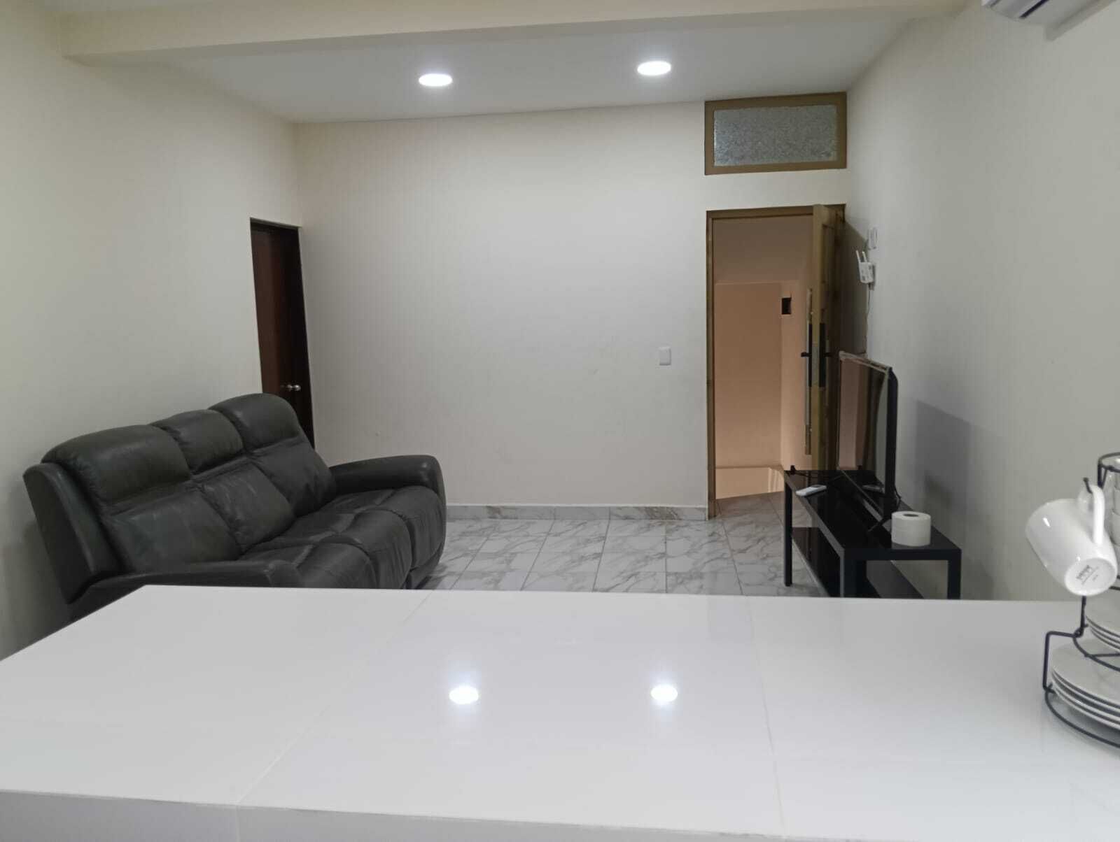 Una habitación con una mesa blanca, un sofá de cuero, un mueble para televisión con un control remoto y una puerta que da a otra habitación.