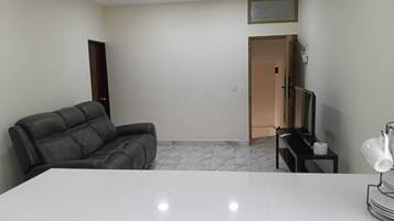 Una habitación con una mesa blanca, un sofá de cuero, un mueble para televisión con un control remoto y una puerta que da a otra habitación.