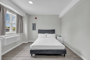 A modern bedroom with a bed, a nightstand, a window with curtains, a wall-mounted control panel, and a ceiling light.
