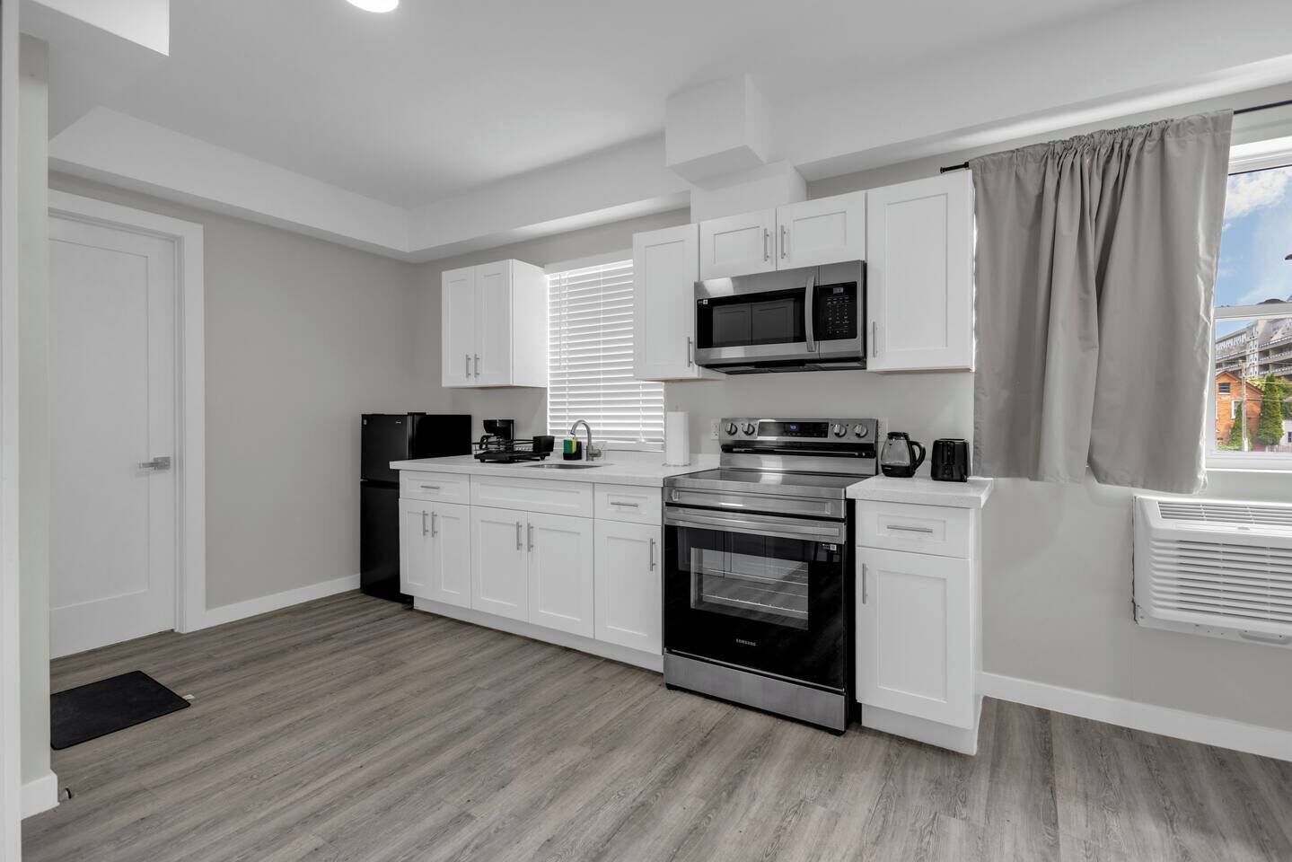Una cocina moderna con armarios blancos, microondas de acero inoxidable, horno empotrado y ventana con persianas.