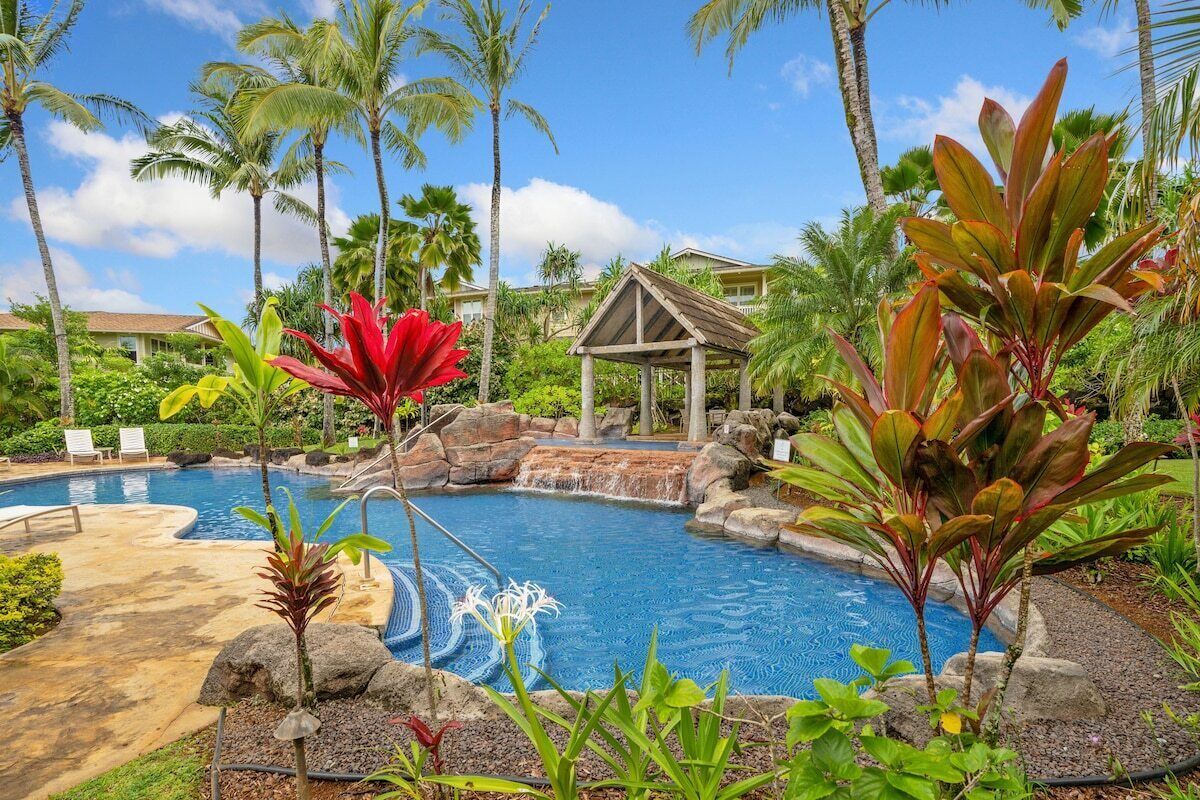 A swimming pool with a waterfall feature, surrounded by tropical plants and palm trees.