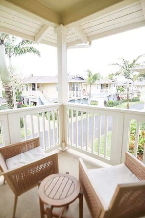 A covered outdoor seating area with wicker furniture and a round table, overlooking a residential street with houses and palm trees. - Modern Island Retreat | Pool & A/C | Mins to Beach (Princeville)