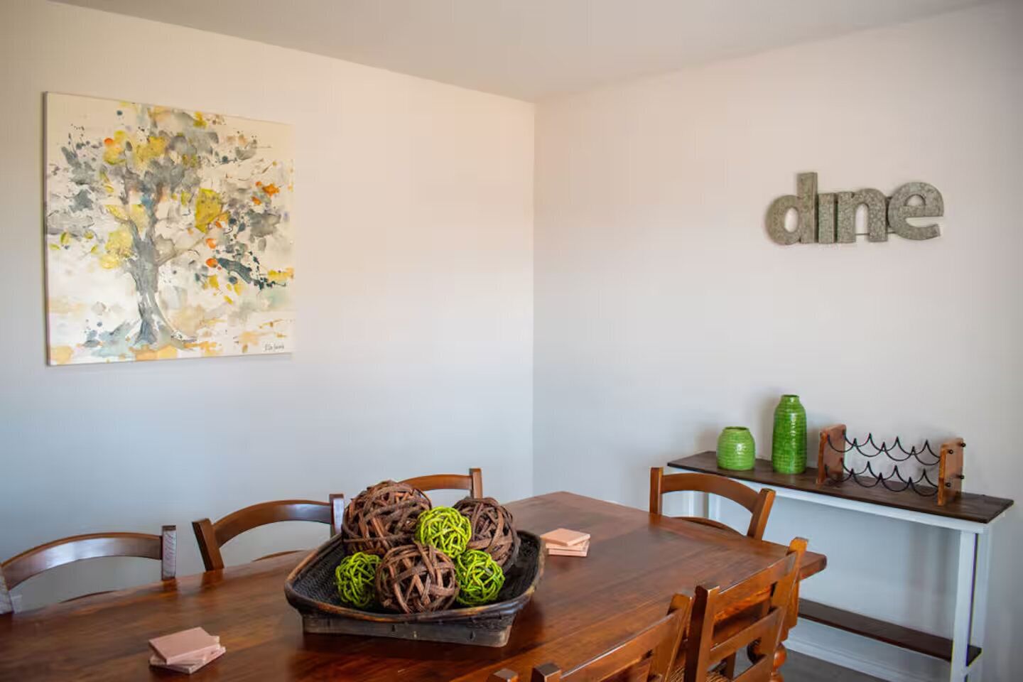 A dining area with a wooden table, chairs, a decorative centerpiece, and a wall-mounted word art piece.