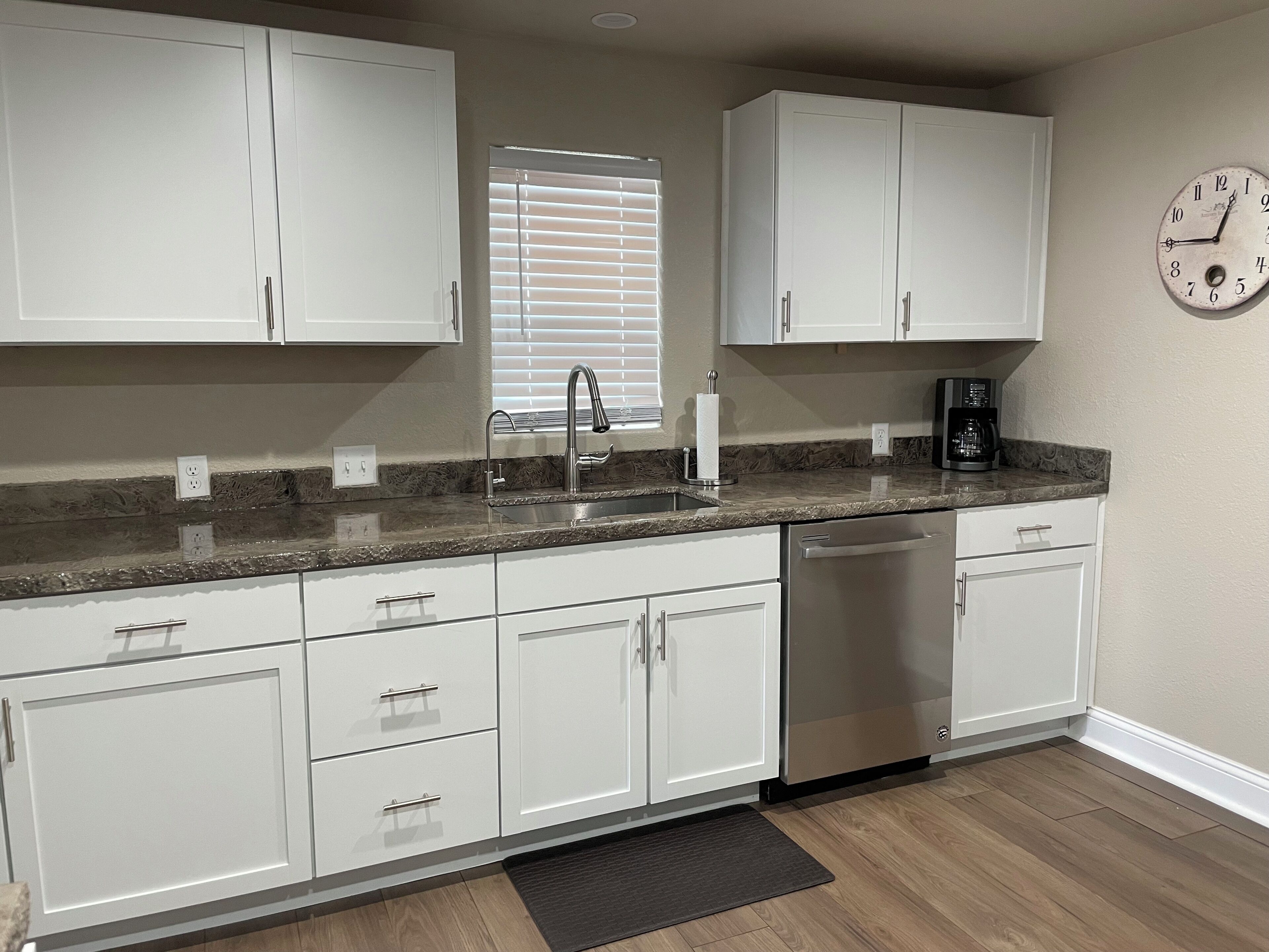 A kitchen with white cabinets, a stainless steel sink, a coffee maker, and a clock on the wall.