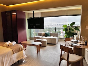 Een moderne hotelkamer met een bank, een eettafel gedekt voor twee personen, een aan de muur bevestigde televisie en een balkon met uitzicht over de stad.