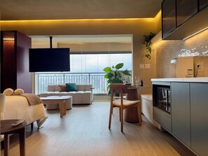 Un salon moderne avec un canapé, une table basse et un téléviseur. On y trouve également un coin cuisine avec des placards et une plante.
