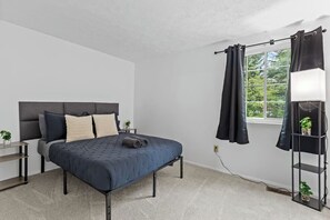 Une chambre à coucher avec un lit, une table de chevet, une fenêtre avec des rideaux et une plante posée sur une étagère.