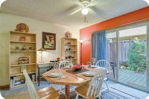 Un coin repas avec une table dressée pour quatre personnes, entourée de chaises blanches, d’un ventilateur de plafond et donnant sur un jardin par une porte-fenêtre coulissante.