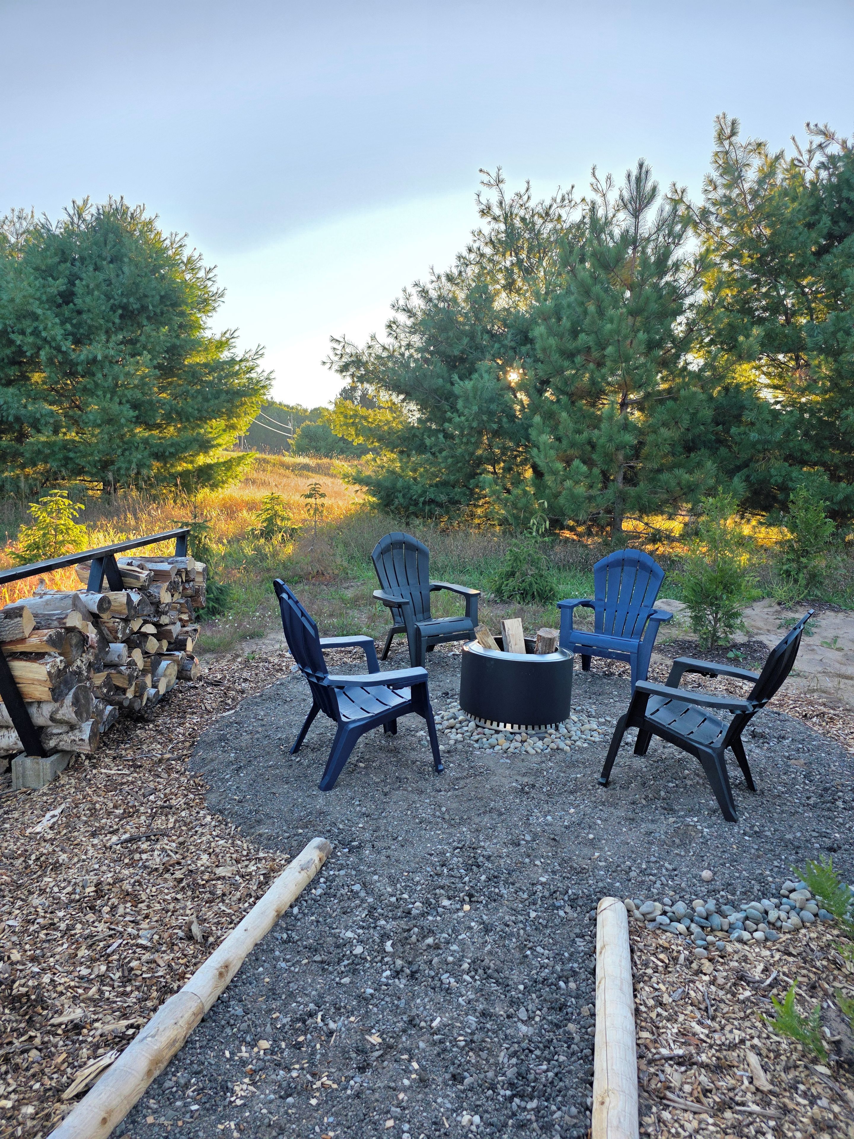 A seating area with four chairs and a central fire pit, surrounded by a gravel ground and a stack of firewood.