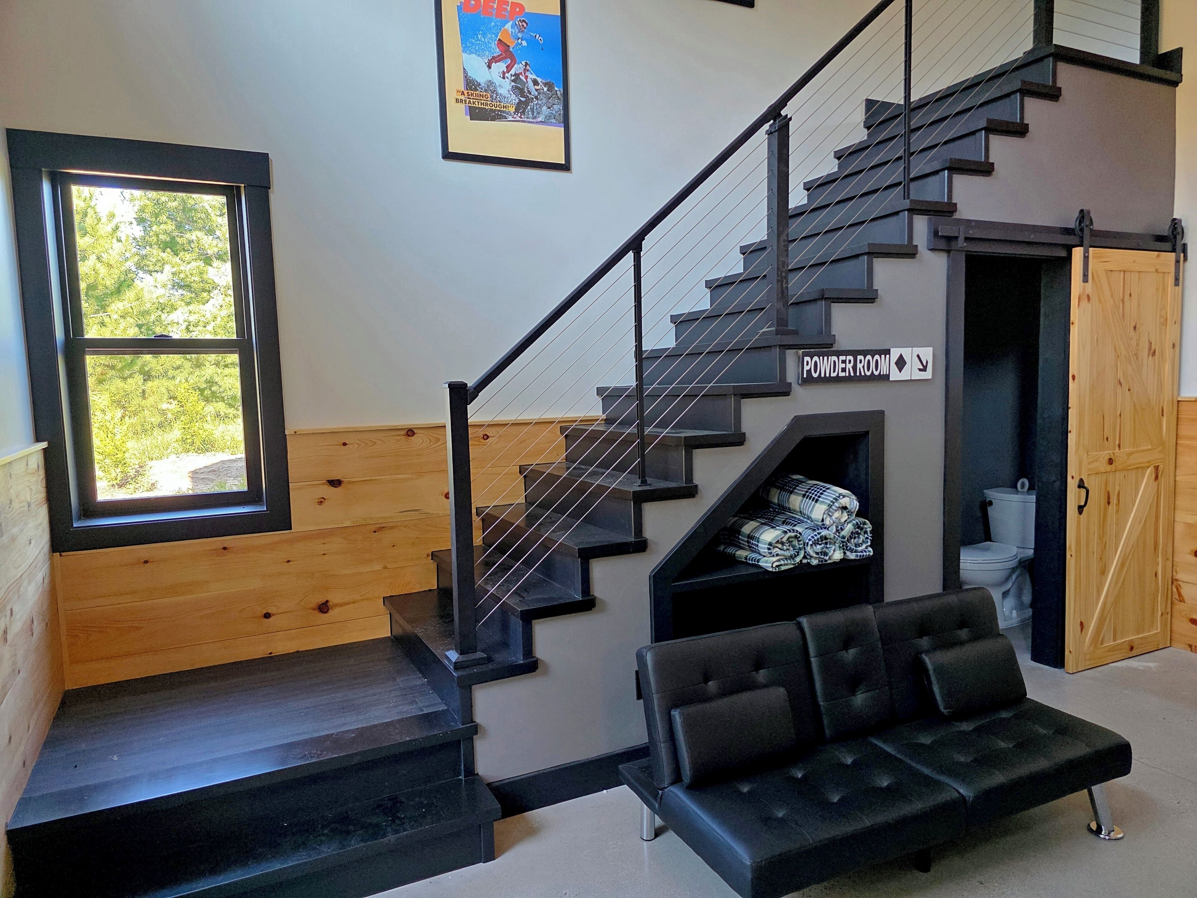Eine Treppe mit Hinweis auf eine Toilette, ein schwarzes Sofa und ein Fenster mit Blick auf Grünflächen.
