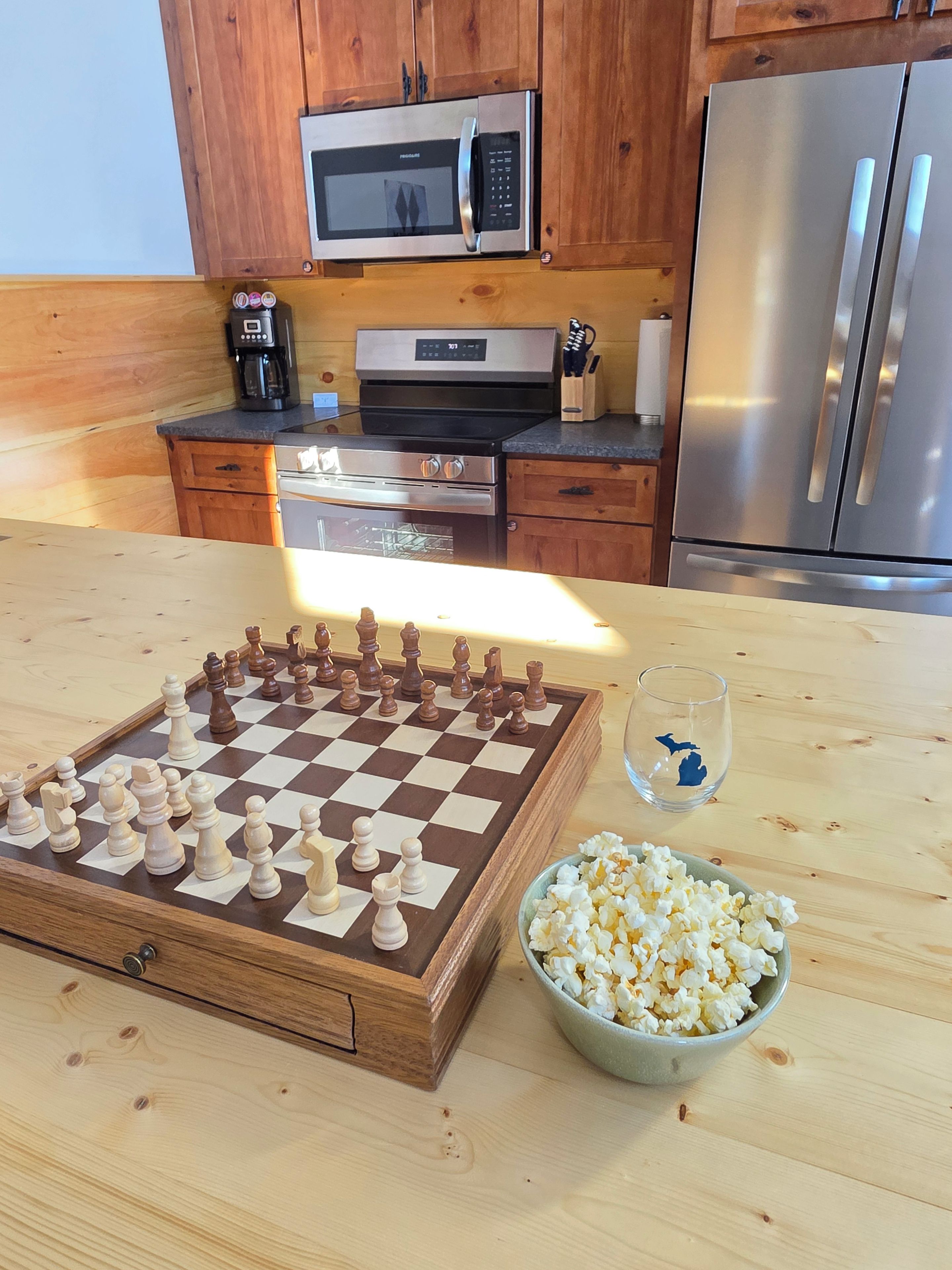 Eine Küche mit Holzschränken, einer Mikrowelle, einer Kaffeemaschine, einem Kühlschrank, einem Schachbrett und einer Schüssel Popcorn.