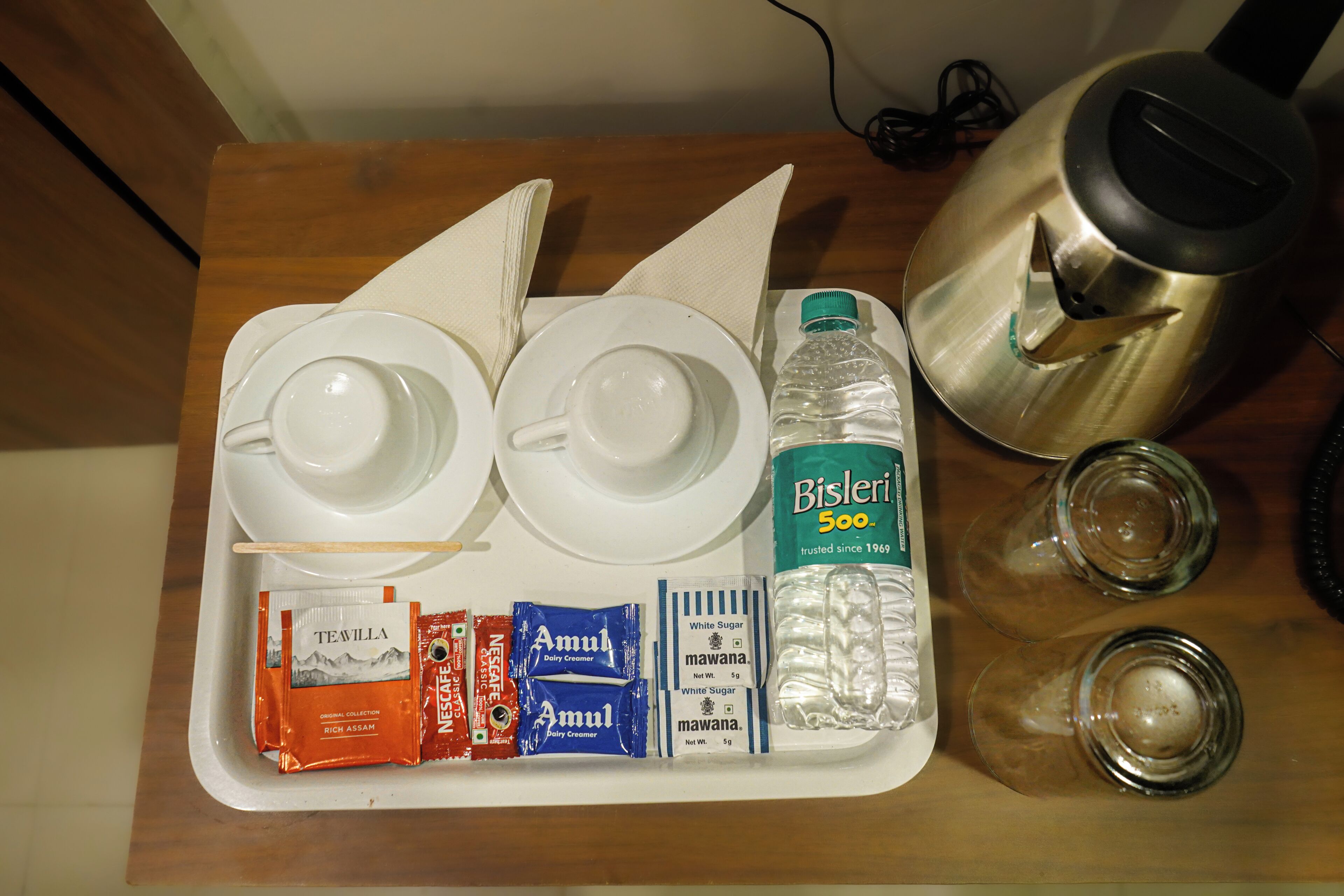 A tray with a teapot, two cups, a bottle of Bisleri water, and packets of tea and cream.