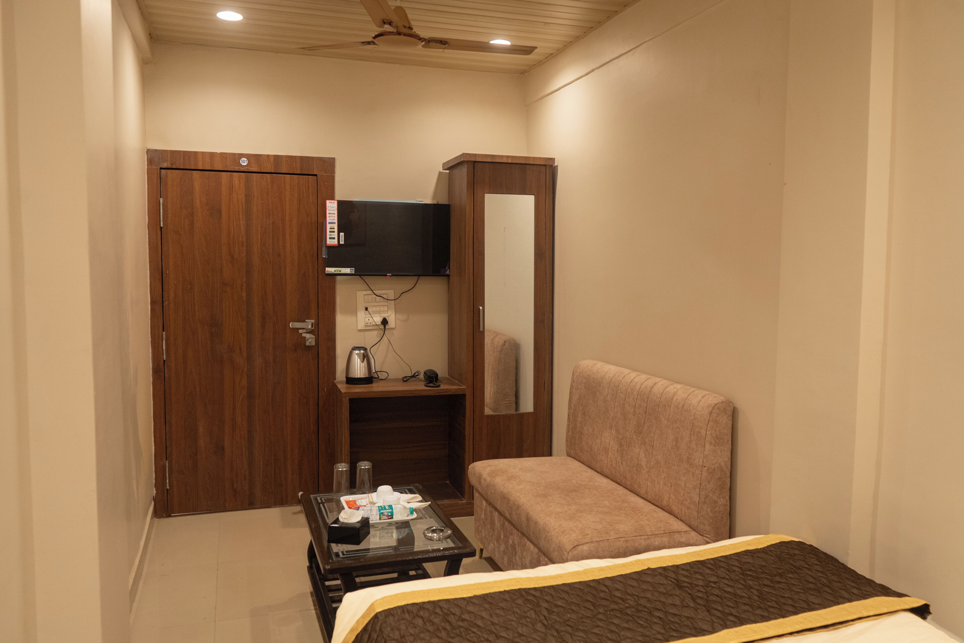 A compact hotel room with a single bed, a small table with a kettle and cups, a television mounted on the wall, and a wooden wardrobe.